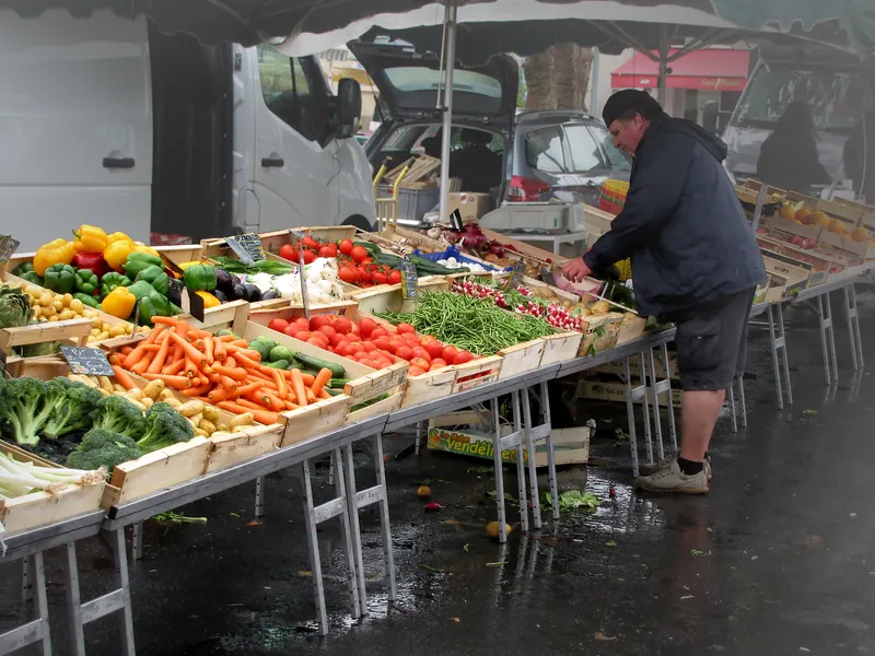 Un maraîcher devant son étal de fruits et légumes sur un marché en plein air, par un matin gris